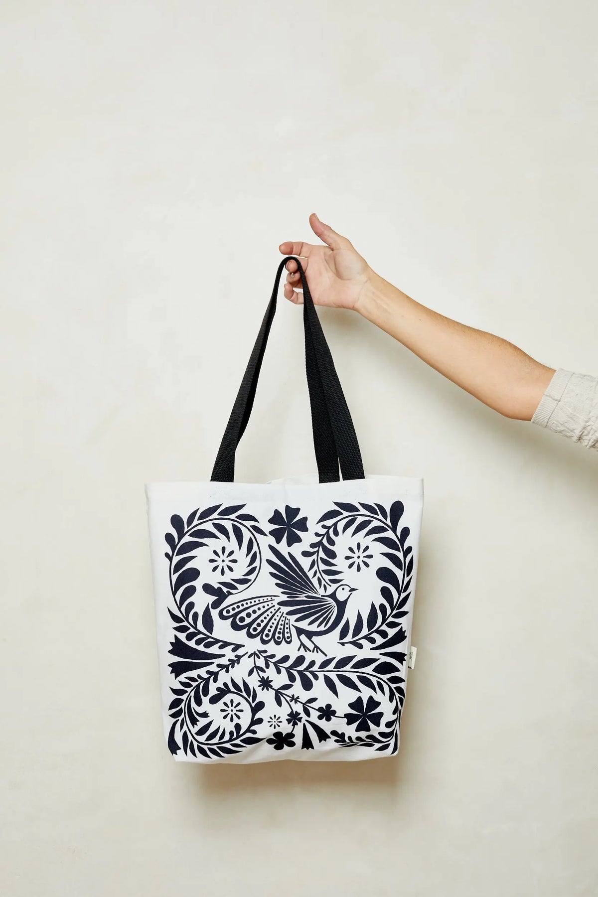 Tote bag de tela blanca con estampado floral y un ave en color azul marino oscuro y asas negras.