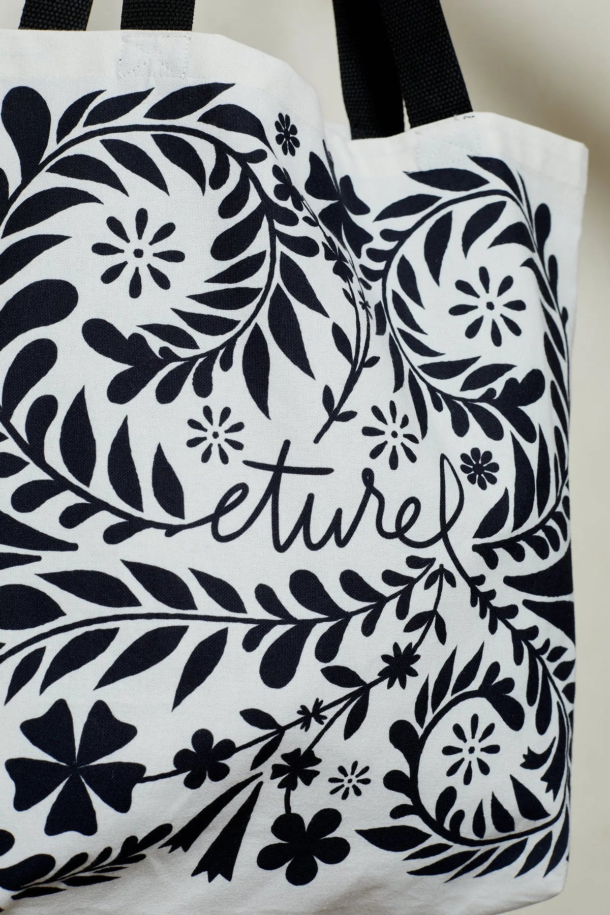 Tote bag de tela blanca con estampado botánico y floral en negro y asas negras. Estilo artesanal.