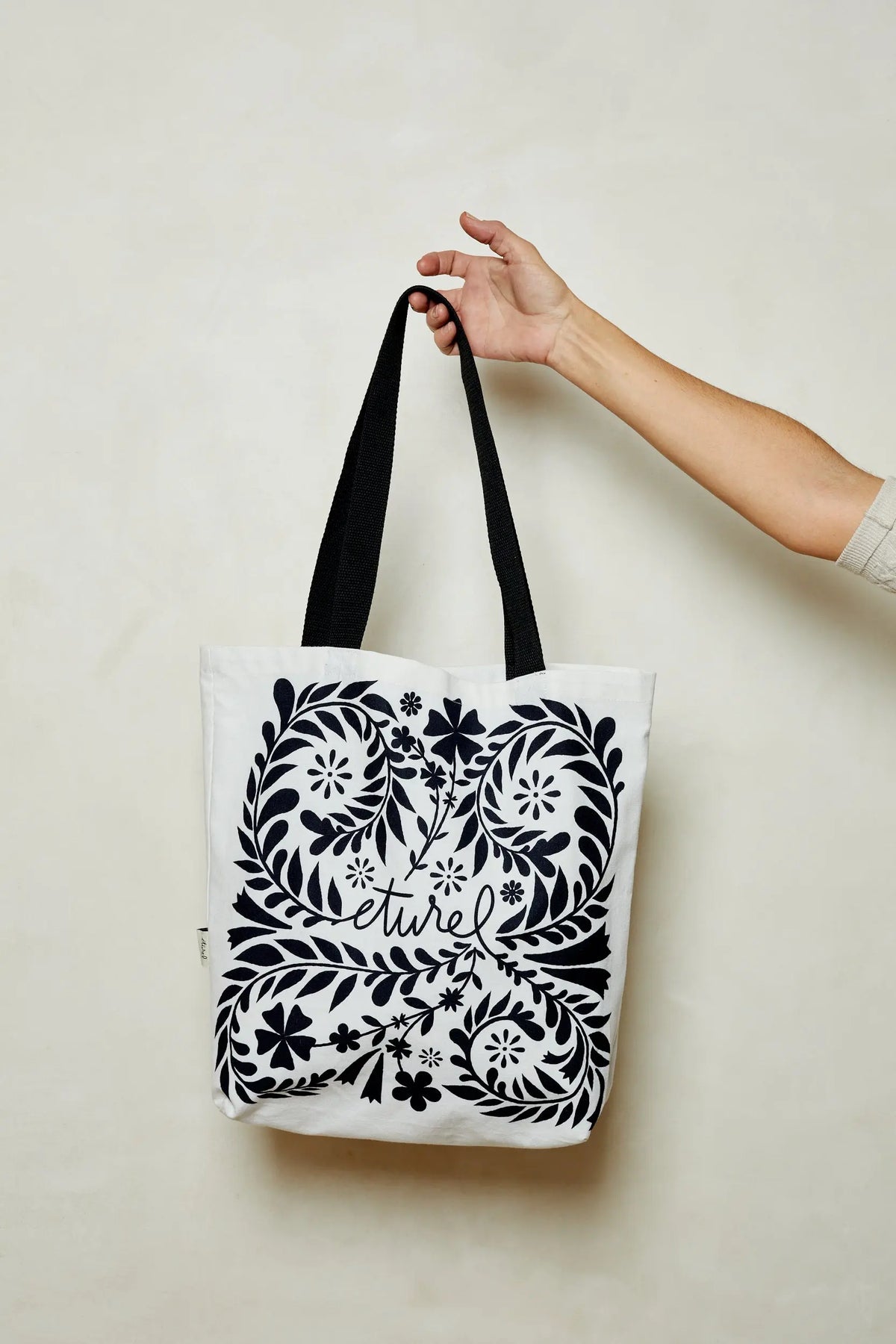Tote bag de tela blanca con asas negras y estampado floral simétrico en color negro. Sostenida por una mano.