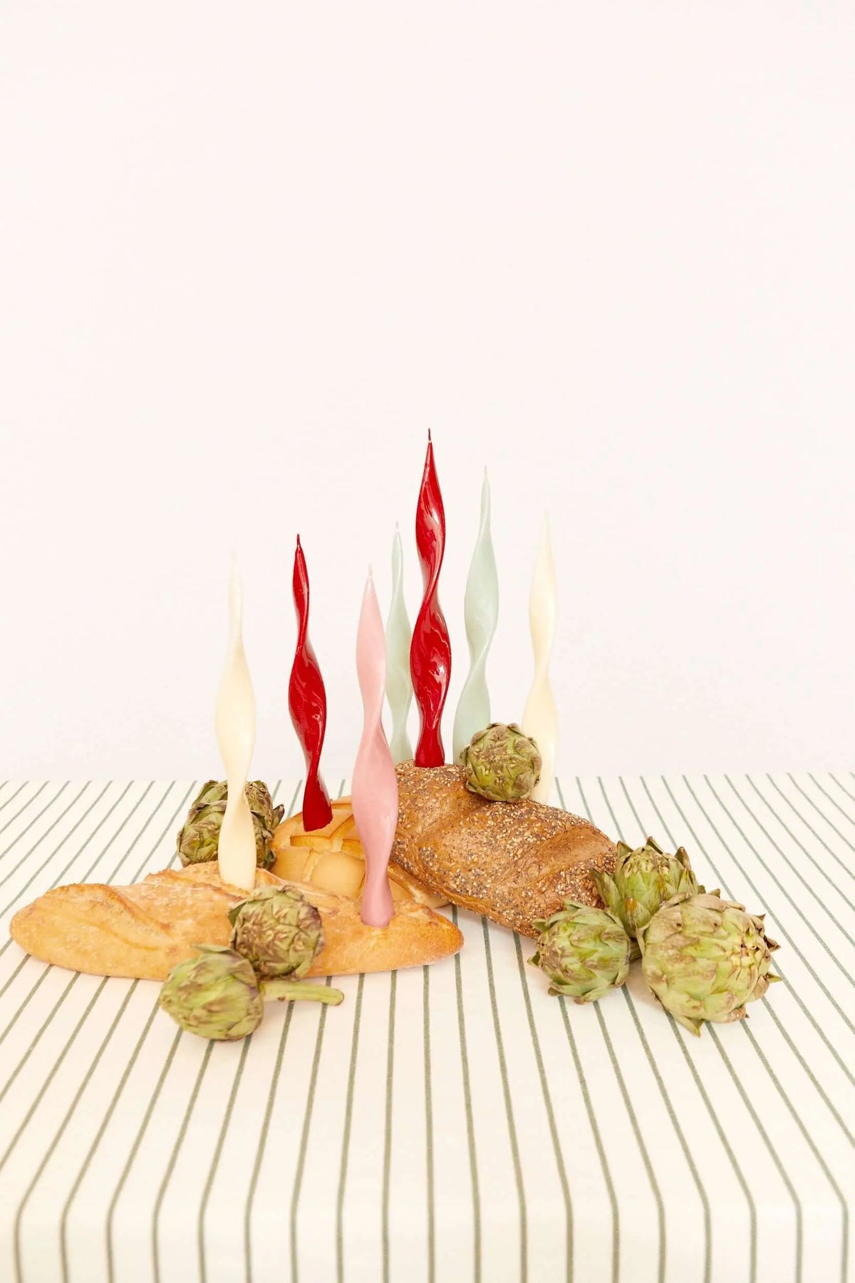Green striped tablecloth with fresh artichokes and bread loaves, topped with colorful, spiral-shaped taper candles.