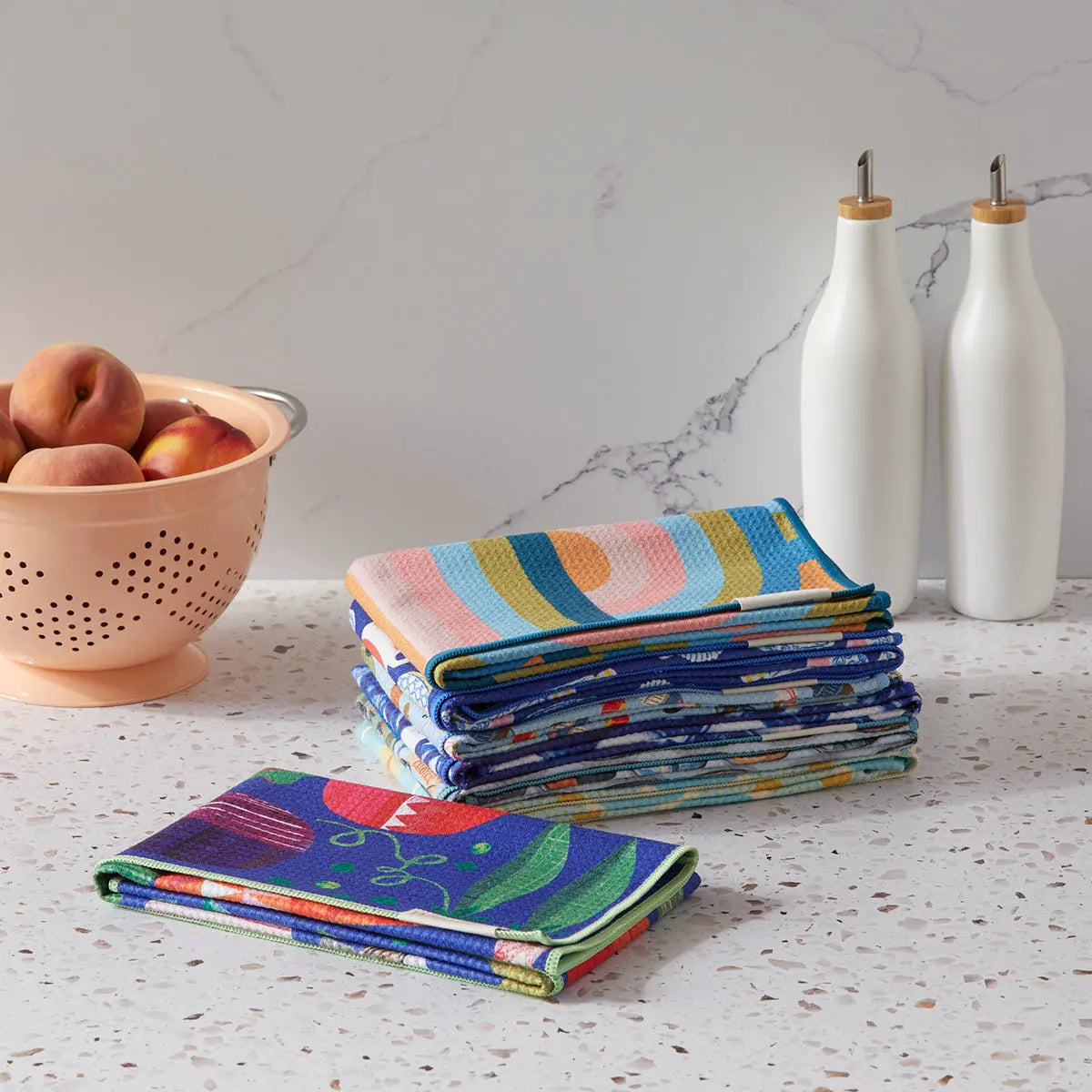 Folded microfiber towels with colorful, whimsical sardine and geometric patterns on a kitchen counter.