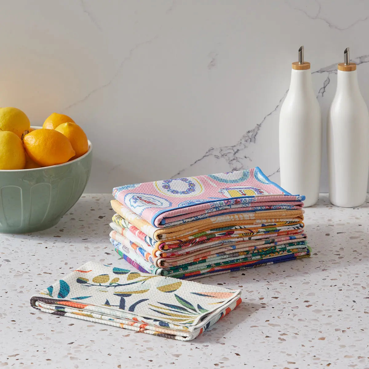 A stack and a single folded Piece of Cake microfiber towel with colorful, decorative patterns on a kitchen counter.
