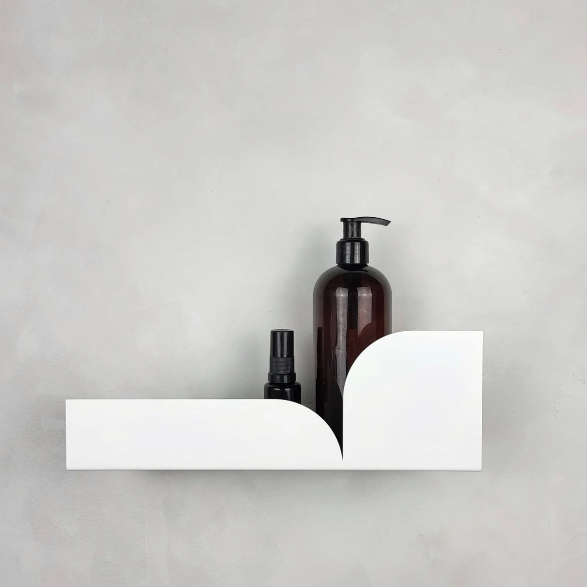 Estante flotante metálico blanco con diseño curvo moderno, sosteniendo dos frascos de baño sobre una pared gris.