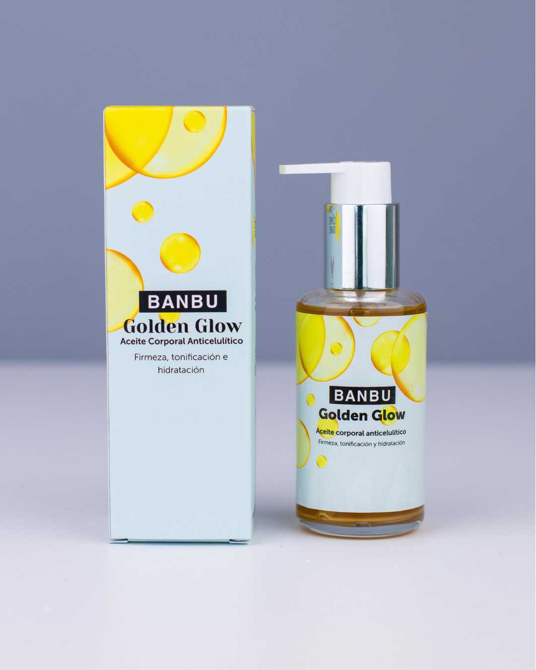 Aceite corporal Golden Glow de Banbu: envase y caja azul claro con gotas doradas sobre fondo gris neutro.