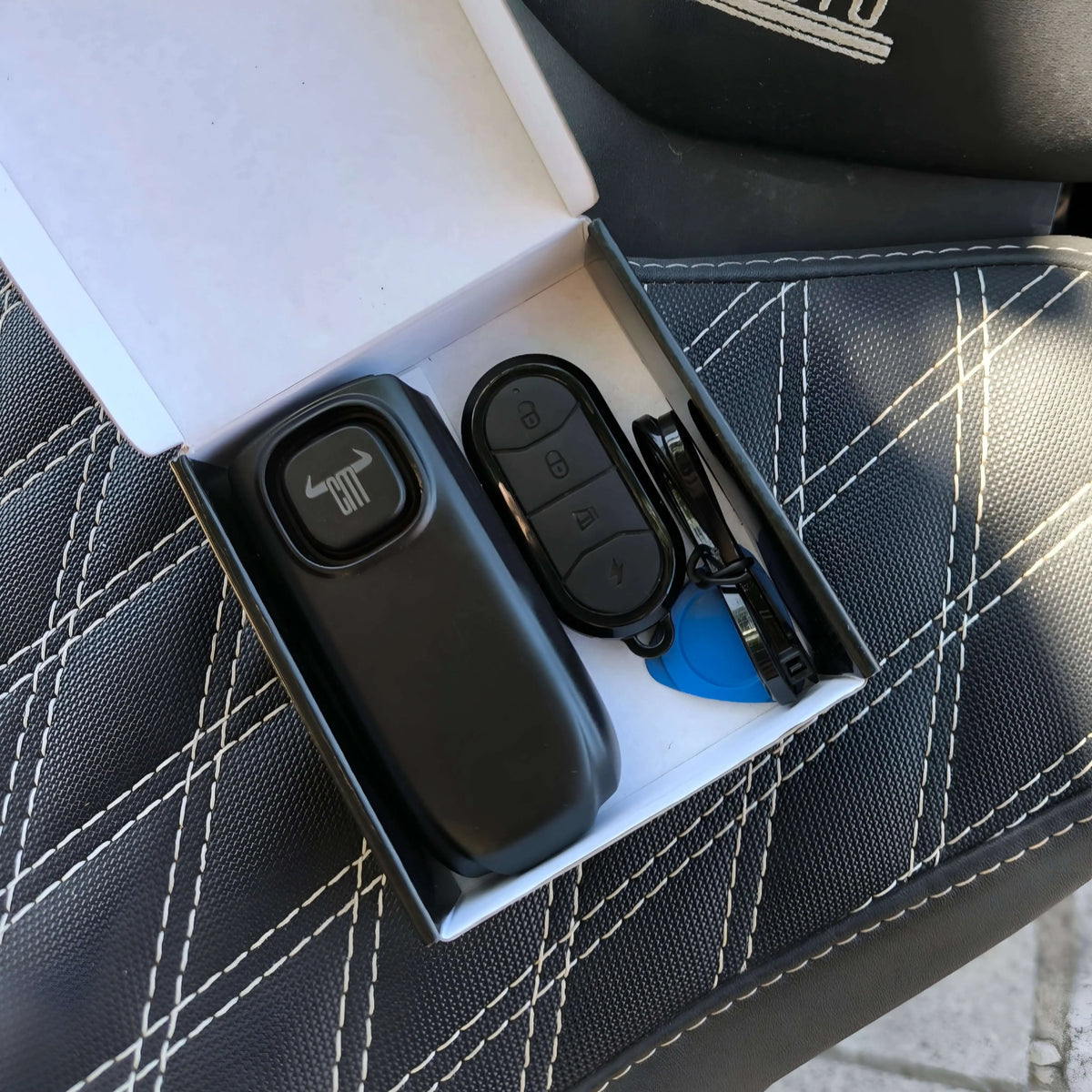 Alarma inalámbrica negra ChuchesMoto con mando y caja sobre asiento de moto con costuras blancas visibles.