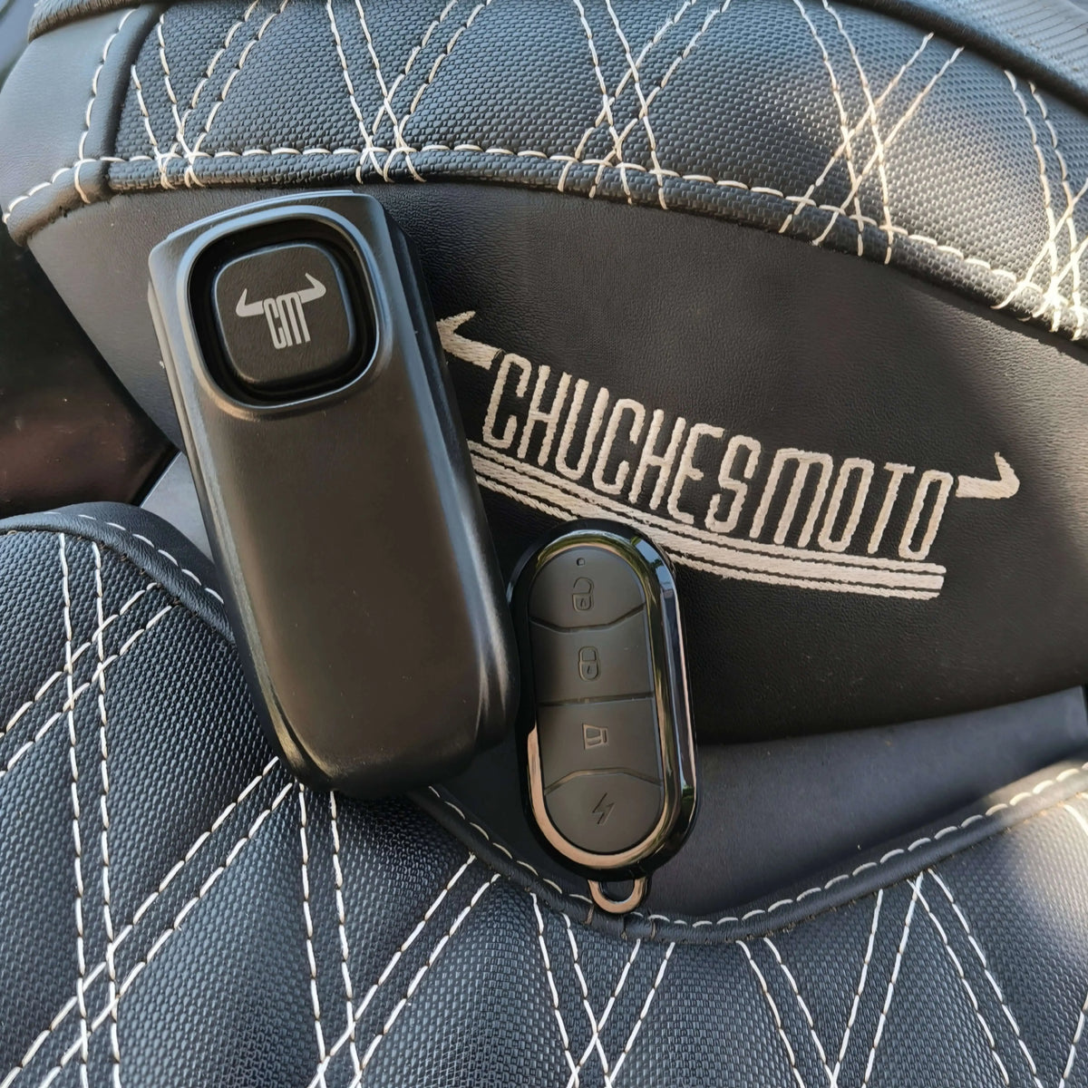 Dos mandos de alarma negros sobre asiento de cuero negro con costuras blancas y logo ChuchesMoto.