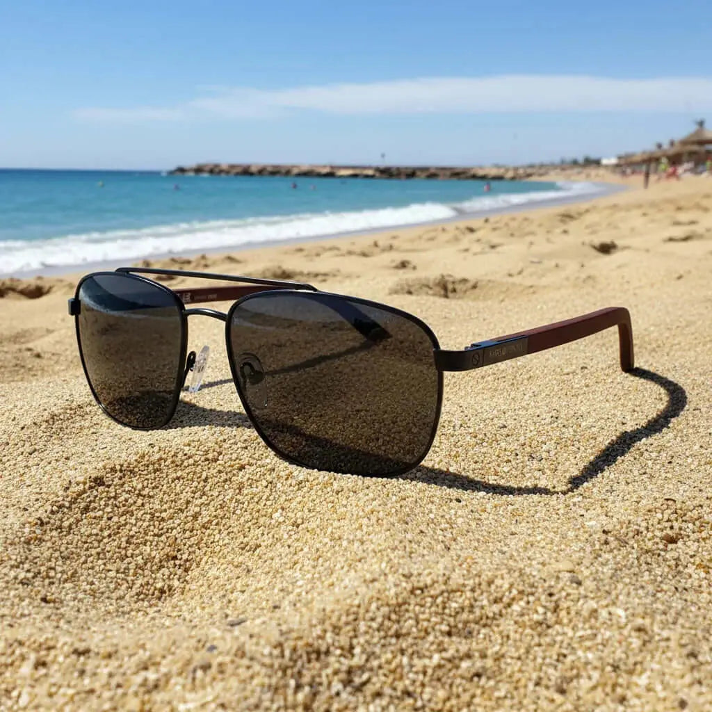 Gafas de sol estilo aviador con montura negra metálica, cristales oscuros y patillas de madera sobre la arena de playa.