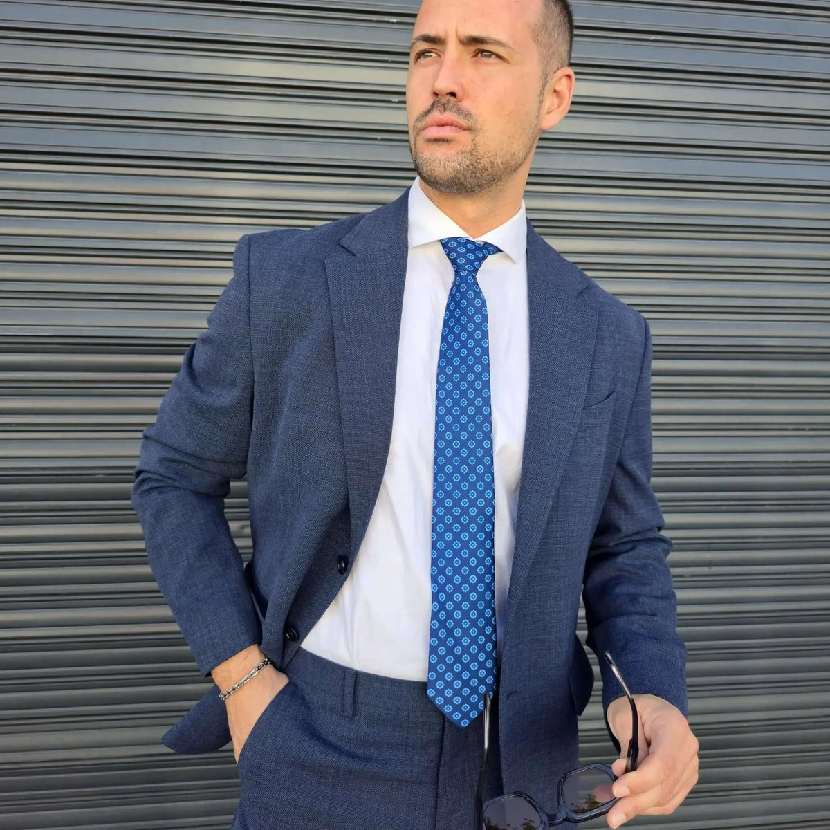 Hombre en traje azul con corbata azul brillante de patrón geométrico, camisa blanca y estilo italiano formal.