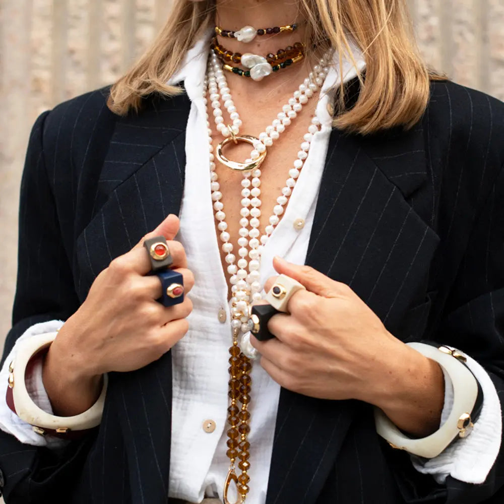 Collares de perlas y ámbar con anillo dorado, blusa blanca, blazer a rayas, y anillos de diferentes estilos en manos.