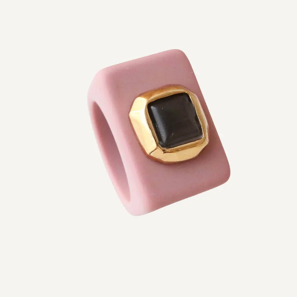 Anillo de silicona rosa con base dorada y piedra negra cuadrada. Diseño moderno y elegante.