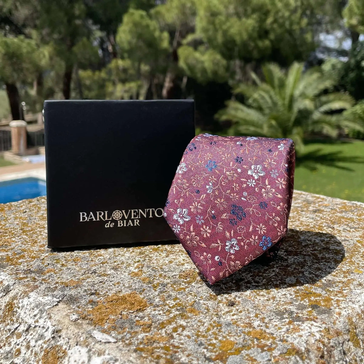 Corbata color granate con estampado floral en tonos azules y dorados, sobre caja negra en un entorno exterior.