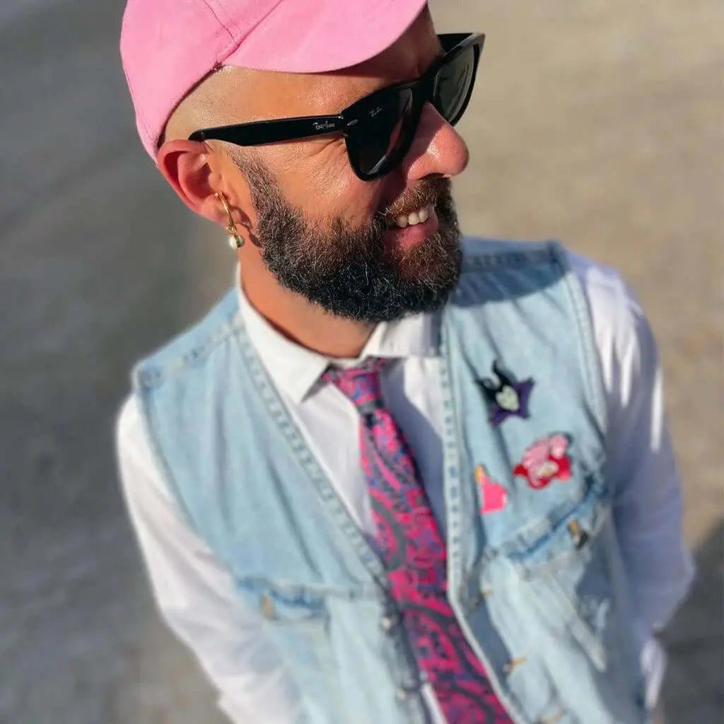 Hombre con gorra rosa, gafas oscuras, chaleco de mezclilla y camisa blanca con corbata de estampado paisley rosa.