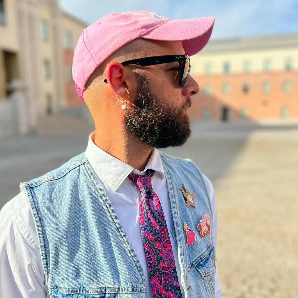 Hombre con gorra rosa, chaleco vaquero, camisa blanca y corbata con estampado paisley vibrante en tonos fucsia y azul.