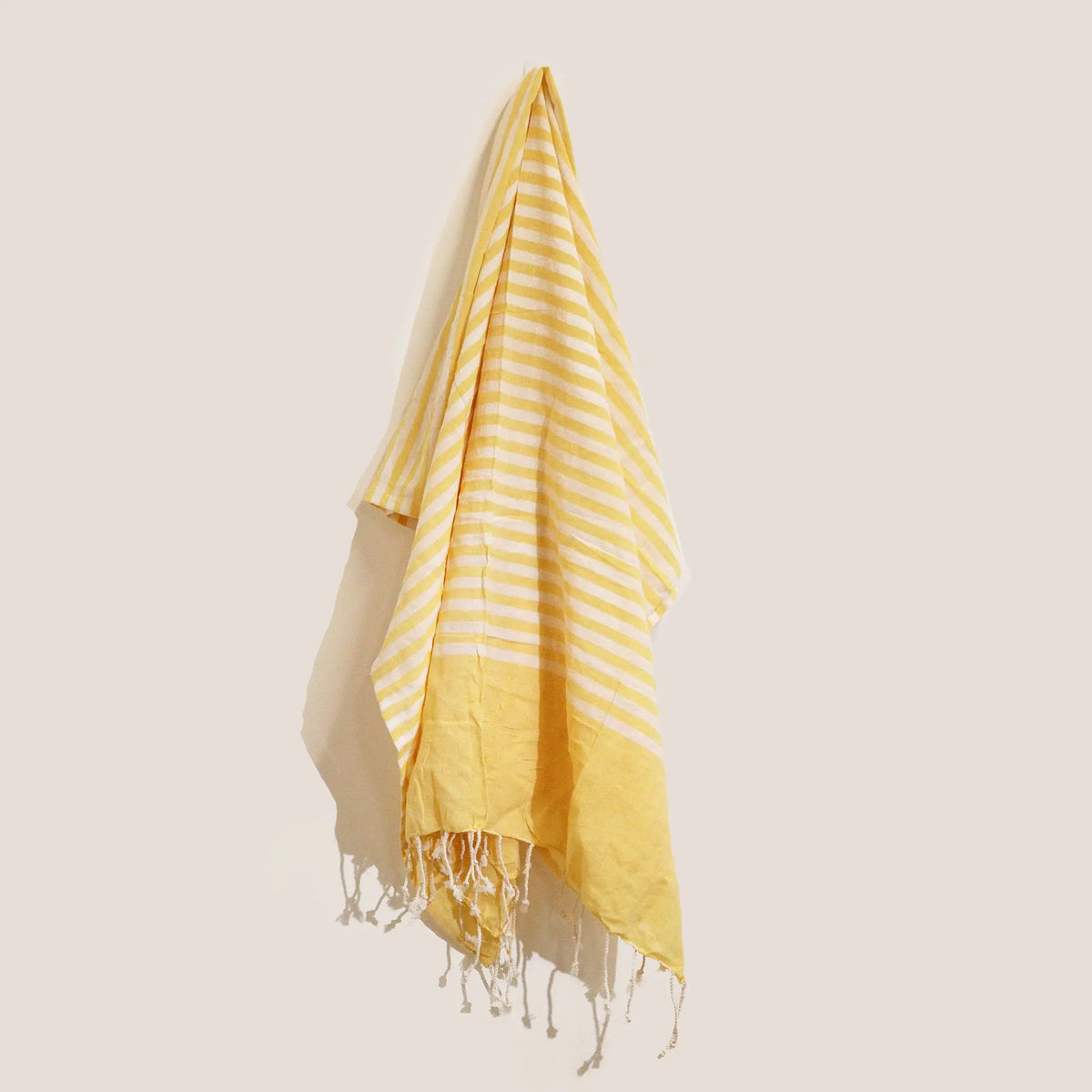 Pareo de algodón amarillo y blanco a rayas con flecos, colgando sobre un fondo neutro.