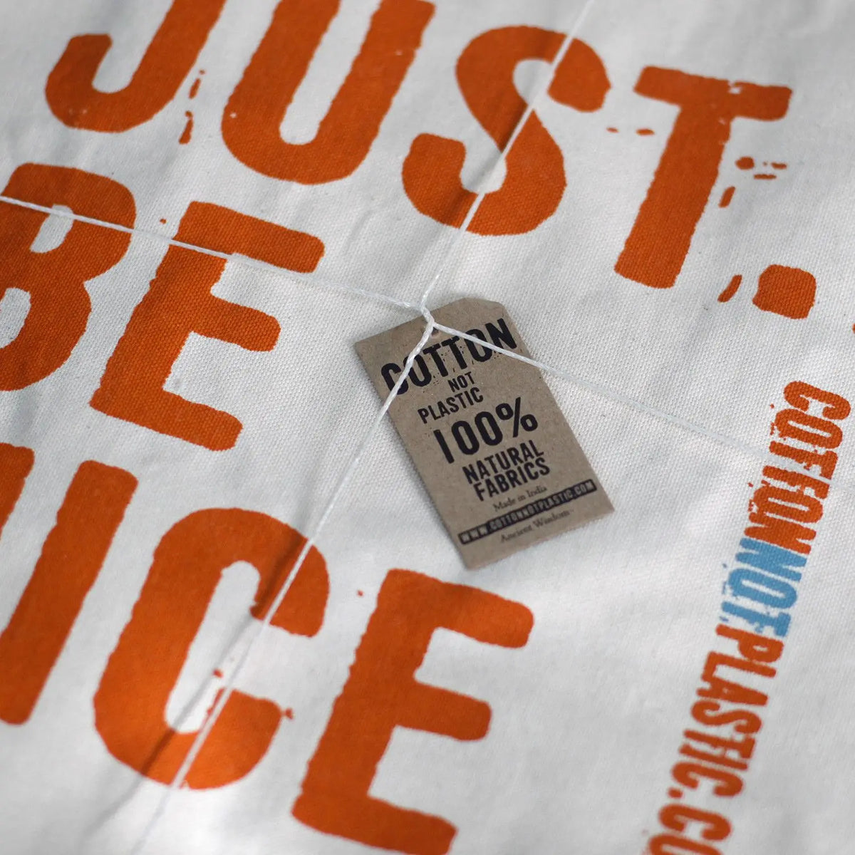 Bolsa de algodón color crema con texto naranja y etiqueta de cartón con información sobre el material 100% natural.