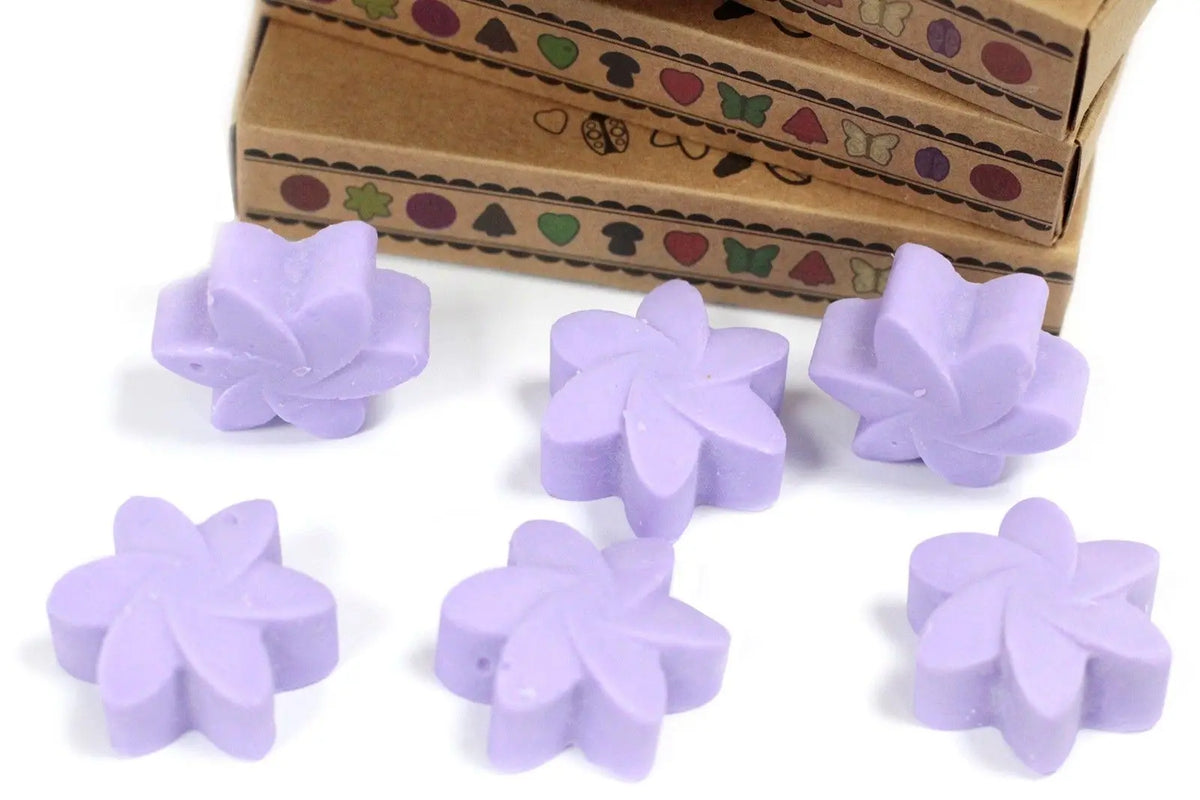 Seis ceras de soja color lavanda con forma de flor frente a cajas de cartón decoradas sobre un fondo blanco.
