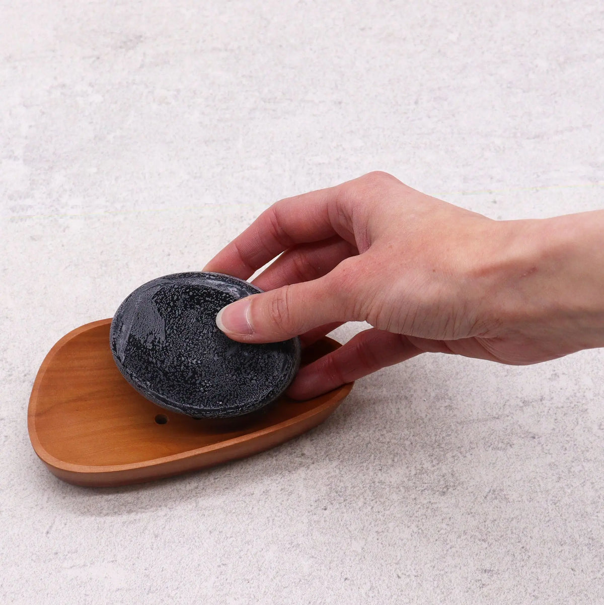 Jabonera de madera de caoba con forma de guijarro, color natural, con pastilla de jabón oscura sostenida por una mano.