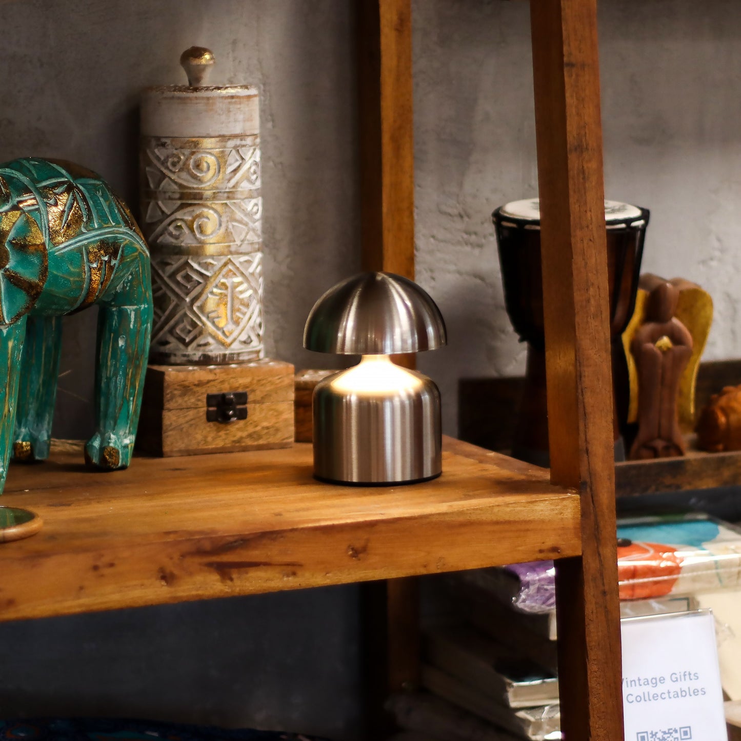 Lámpara de mesa táctil con forma de champiñón, fabricada en metal plateado, emitiendo una luz cálida en una estantería.