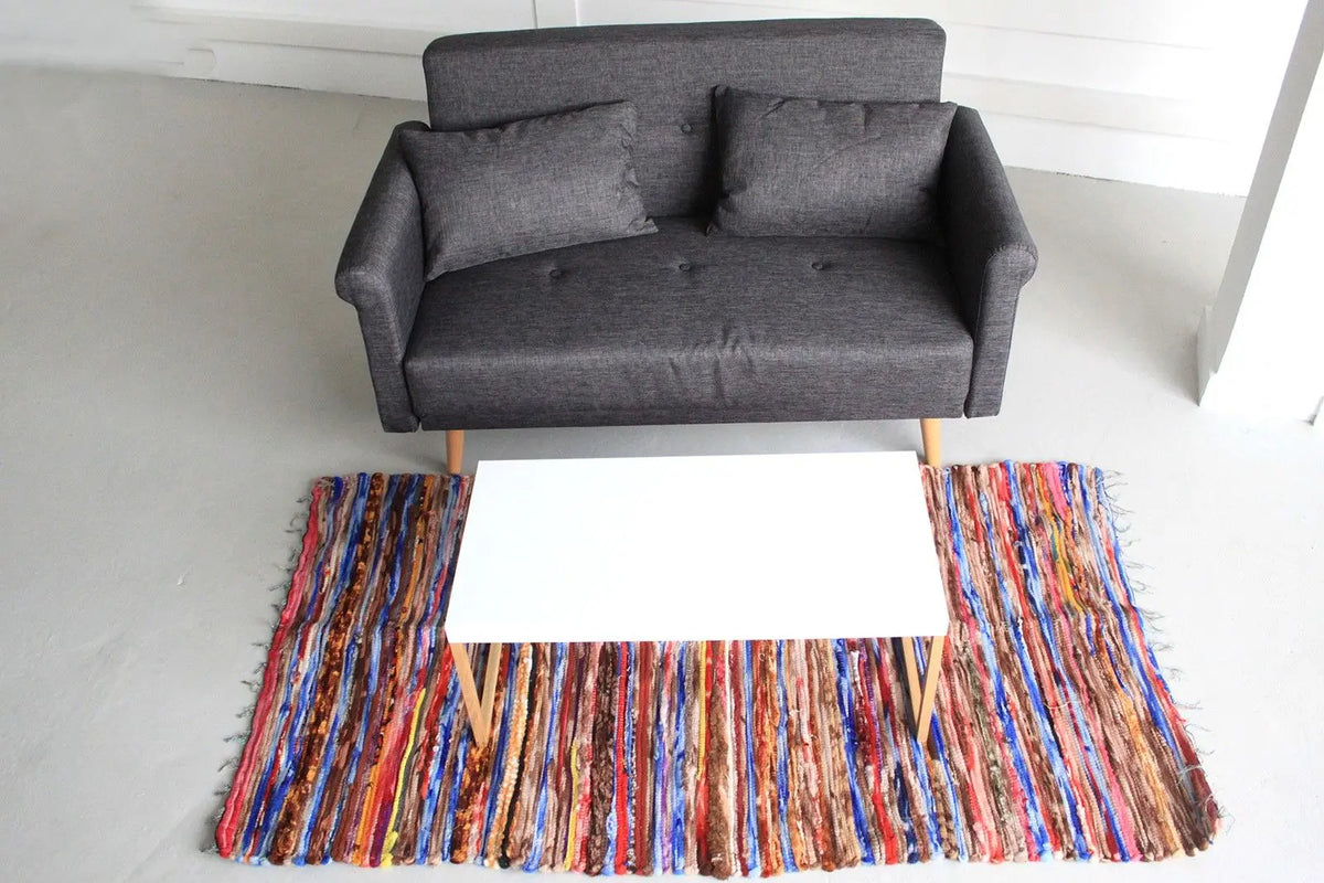 Alfombra de trapo rectangular (105x185cm) con rayas multicolores, tejido artesanal indio frente a un sofá gris.