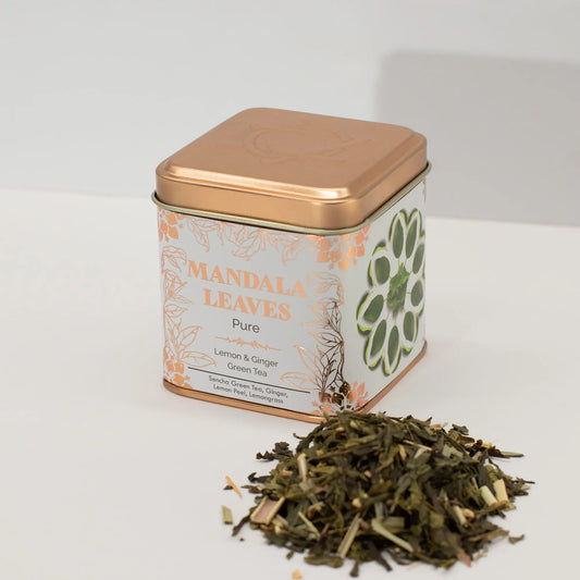 Lata cuadrada color cobre con té verde, limón y jengibre, junto a una muestra de hebras sobre superficie blanca.