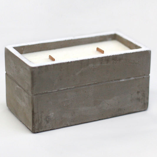 Vela rectangular de cera blanca con dos mechas de madera en un recipiente de hormigón gris con estilo rústico moderno.