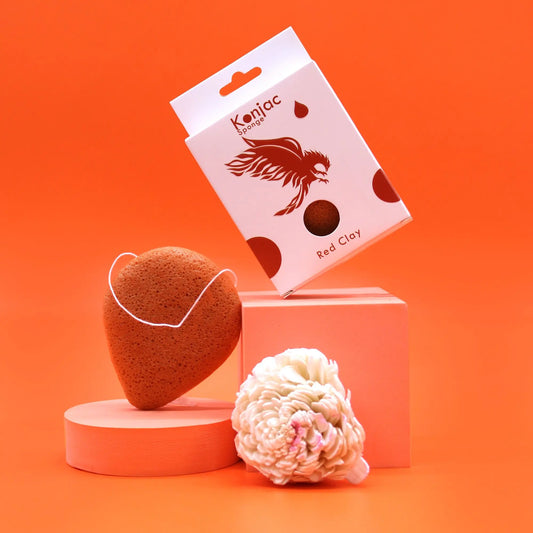 Esponja facial de arcilla roja en forma de gota con cuerda, junto a su caja y una flor decorativa sobre fondo naranja.