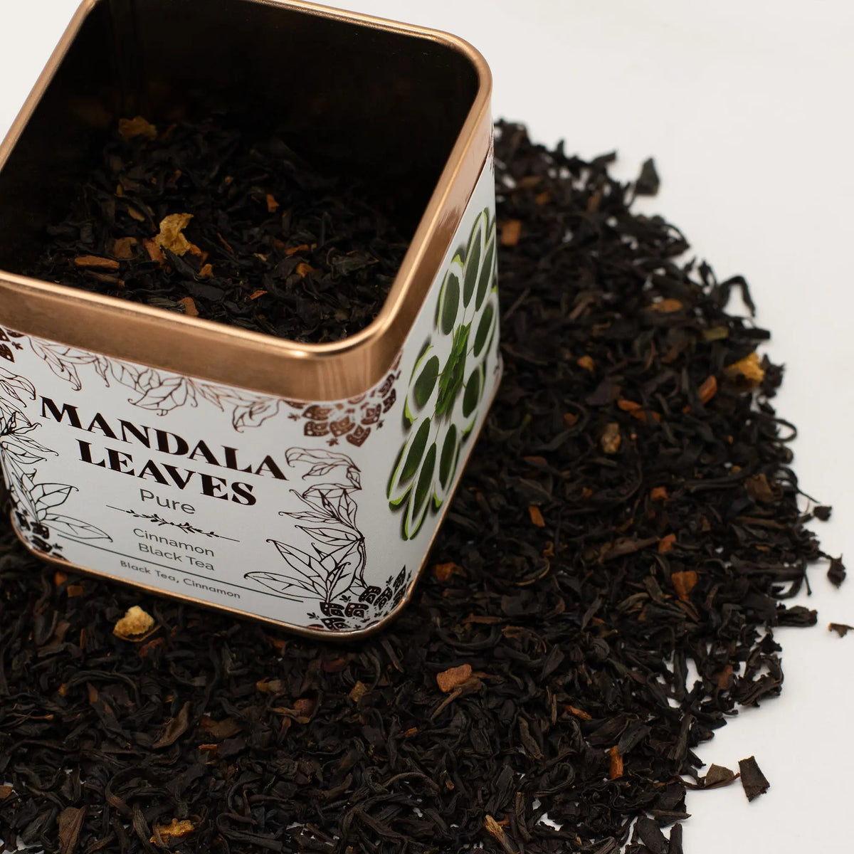 Lata metálica cuadrada abierta con té negro y trozos de fruta sobre un fondo de hojas de té dispersas.