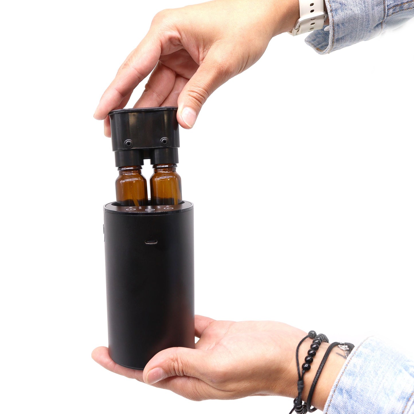 Difusor de aromas negro, cilíndrico, con dos botes de vidrio ámbar y conexión USB-C. Sostenido por manos humanas.