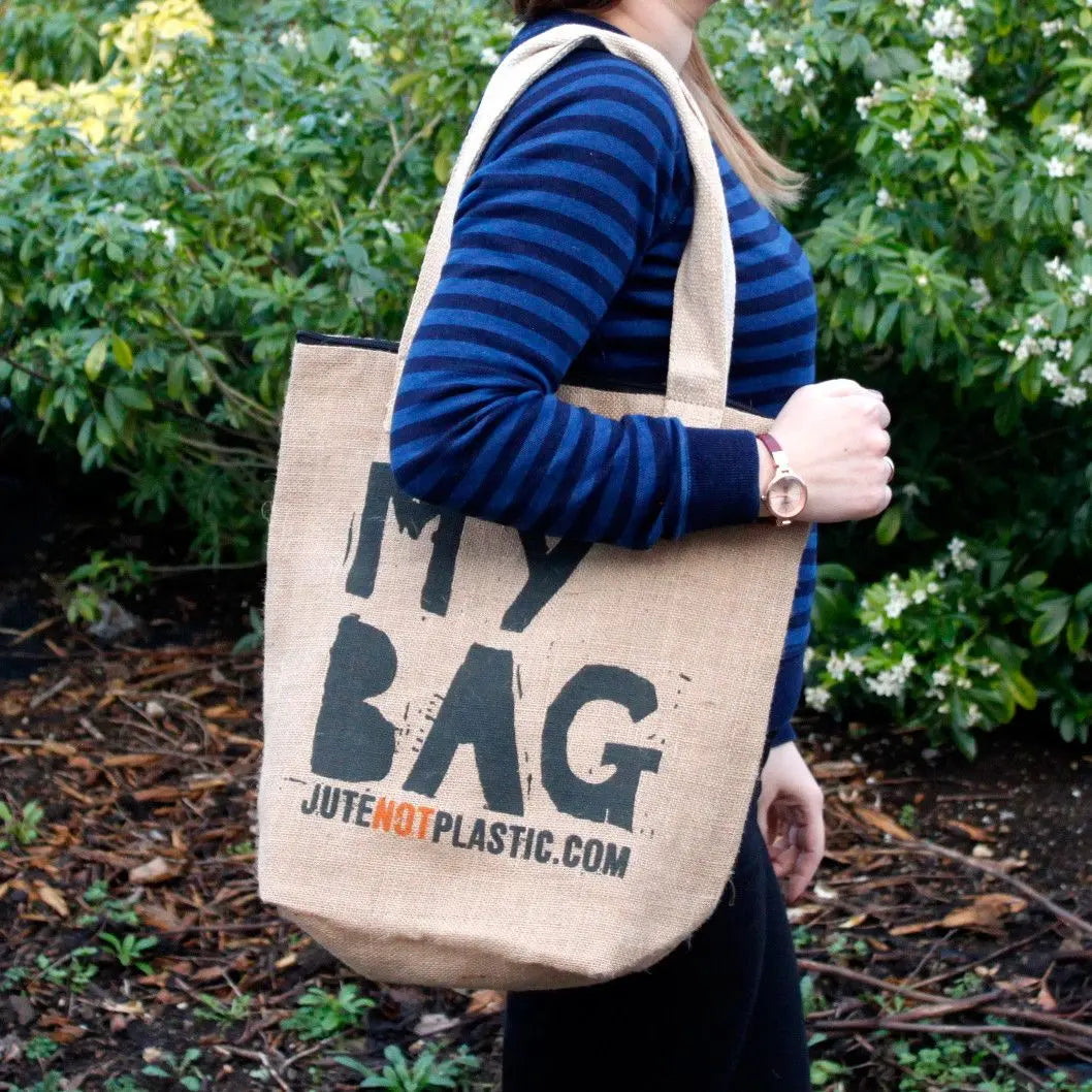 Persona lleva un bolso de hombro de yute color beige con texto negro impreso, ideal para compras ecológicas y casuales.