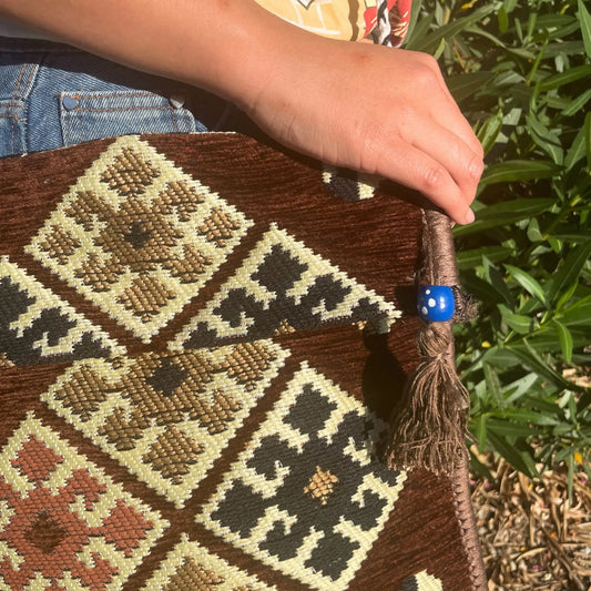 Bolso bandolera de tela tipo kilim, tonos chocolate y crema con rombos, decorado con una cuenta azul y una borla.
