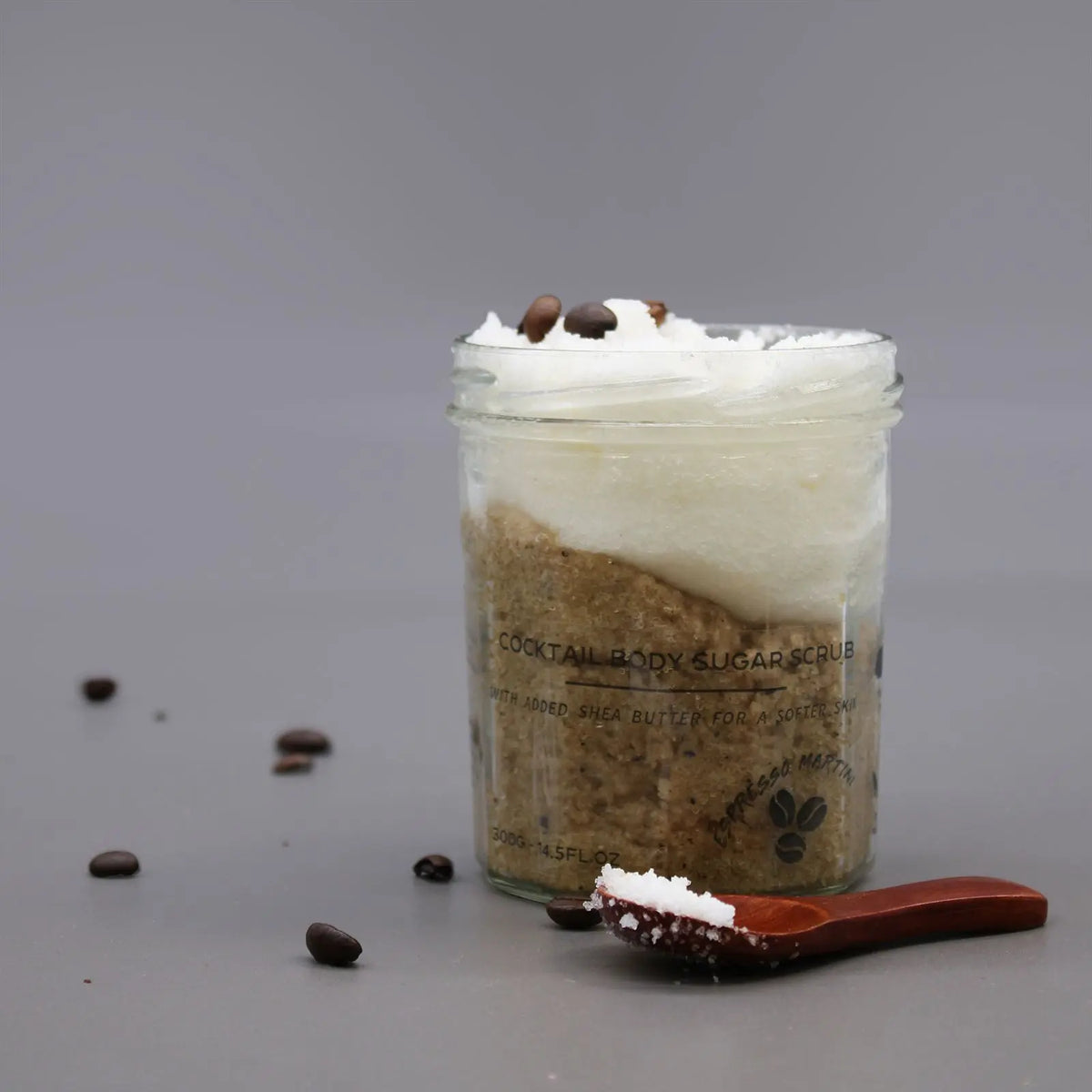Tarro de cristal con exfoliante de azúcar, capa cremosa, granos de café y cuchara de madera sobre fondo gris.