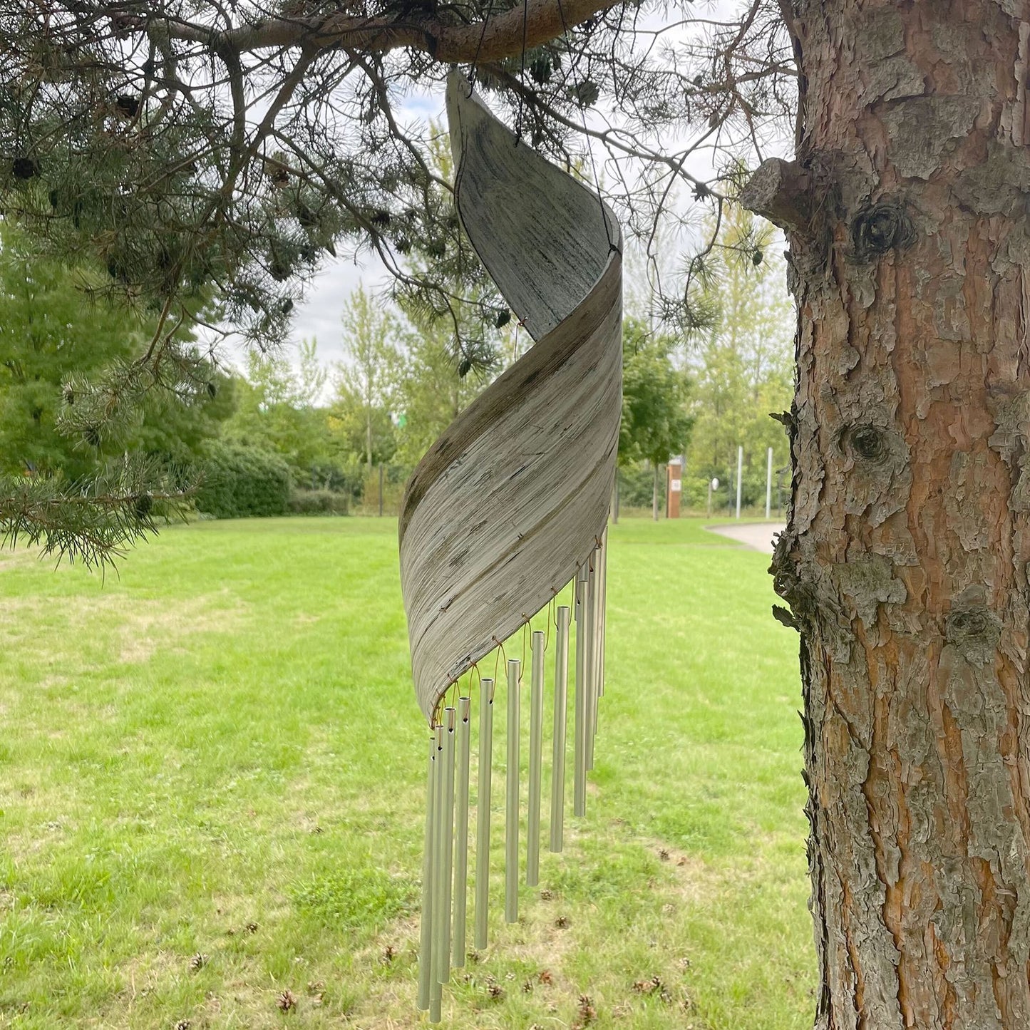 Campanilla de viento en espiral de hoja de coco blanqueada con tubos metálicos colgando de un árbol en un jardín.