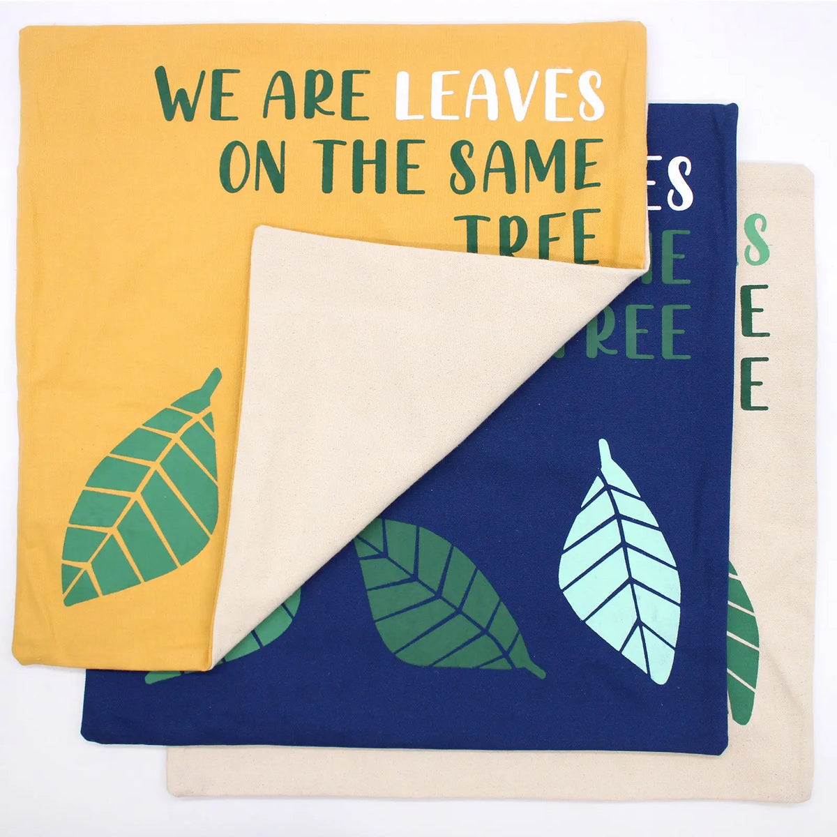 Tres fundas de cojín de algodón con estampado de hojas y texto, en colores amarillo, azul y natural.