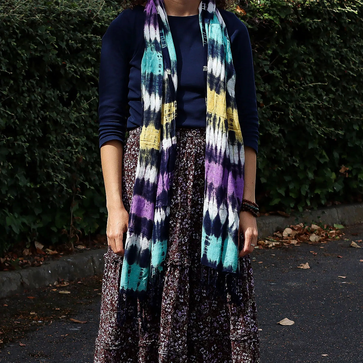 Estola larga estilo boho con rayas horizontales tie-dye en azul, morado, amarillo y blanco, sobre fondo de arbustos.