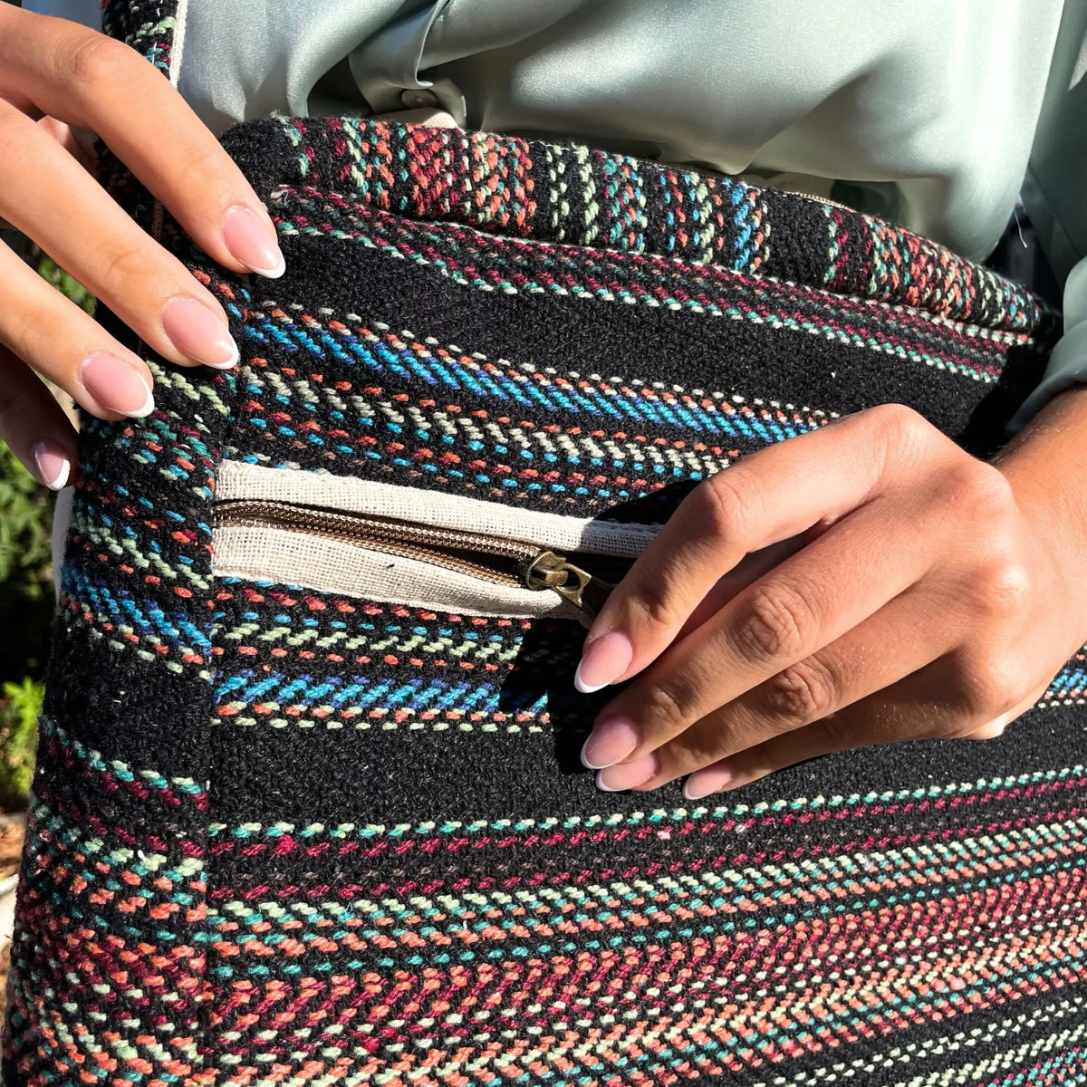 Bolso de tela tejida en negro con rayas multicolores, detalle de cremallera en bolsillo frontal y acabado artesanal.