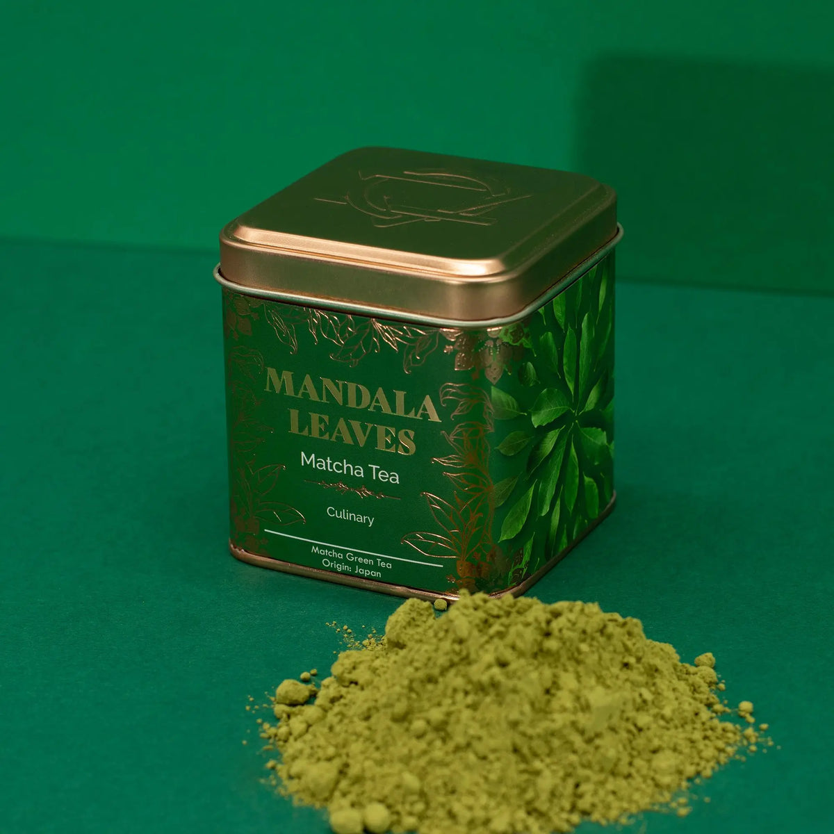 Lata cuadrada verde con detalles dorados y polvo de té matcha frente a ella, sobre un fondo verde uniforme.