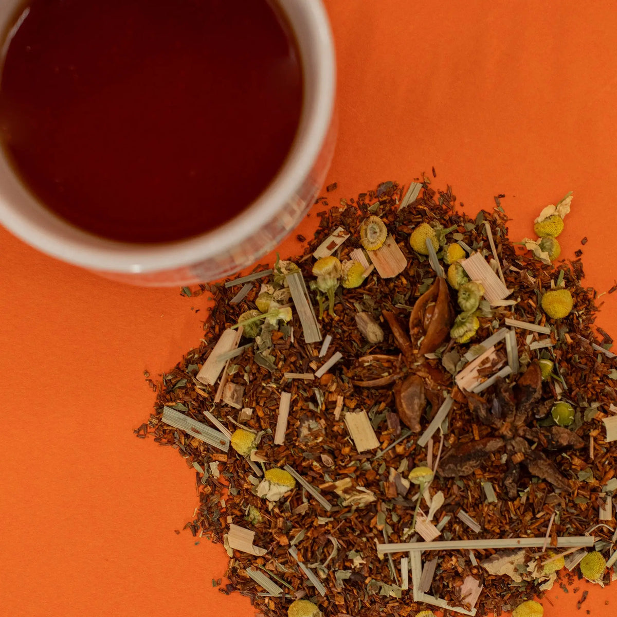 Taza de té rooibos junto a una mezcla de hierbas, flores de manzanilla y anís sobre fondo naranja brillante.
