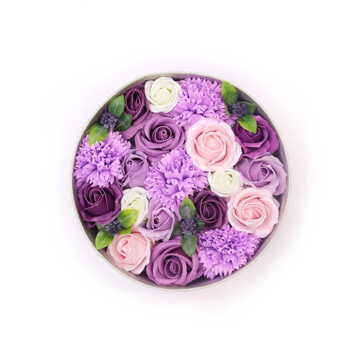 Caja redonda con rosas y claveles de jabón en tonos violeta, lila y blanco con detalles de hojas verdes.