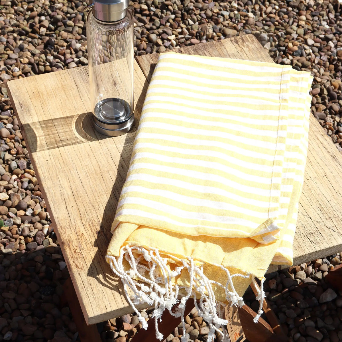 Pareo de algodón amarillo sol con rayas blancas y flecos, sobre mesa de madera rústica al aire libre junto a una botella