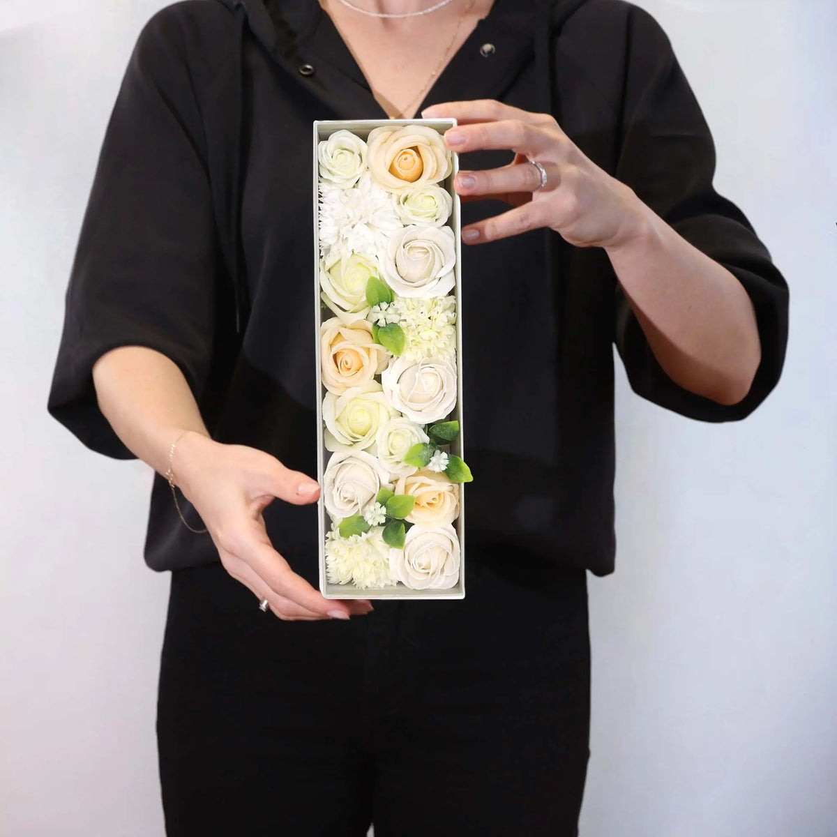 Persona sosteniendo una caja rectangular con rosas decorativas en tonos blanco y marfil con hojas verdes de estilo elegante