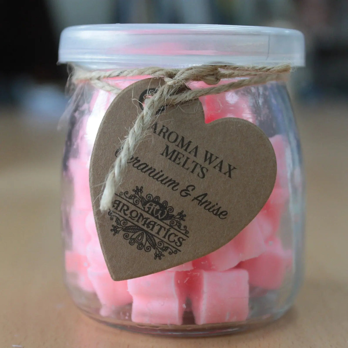 Frasco de vidrio con corazones de cera rosa, etiqueta de cartón en forma de corazón y tapa de plástico.