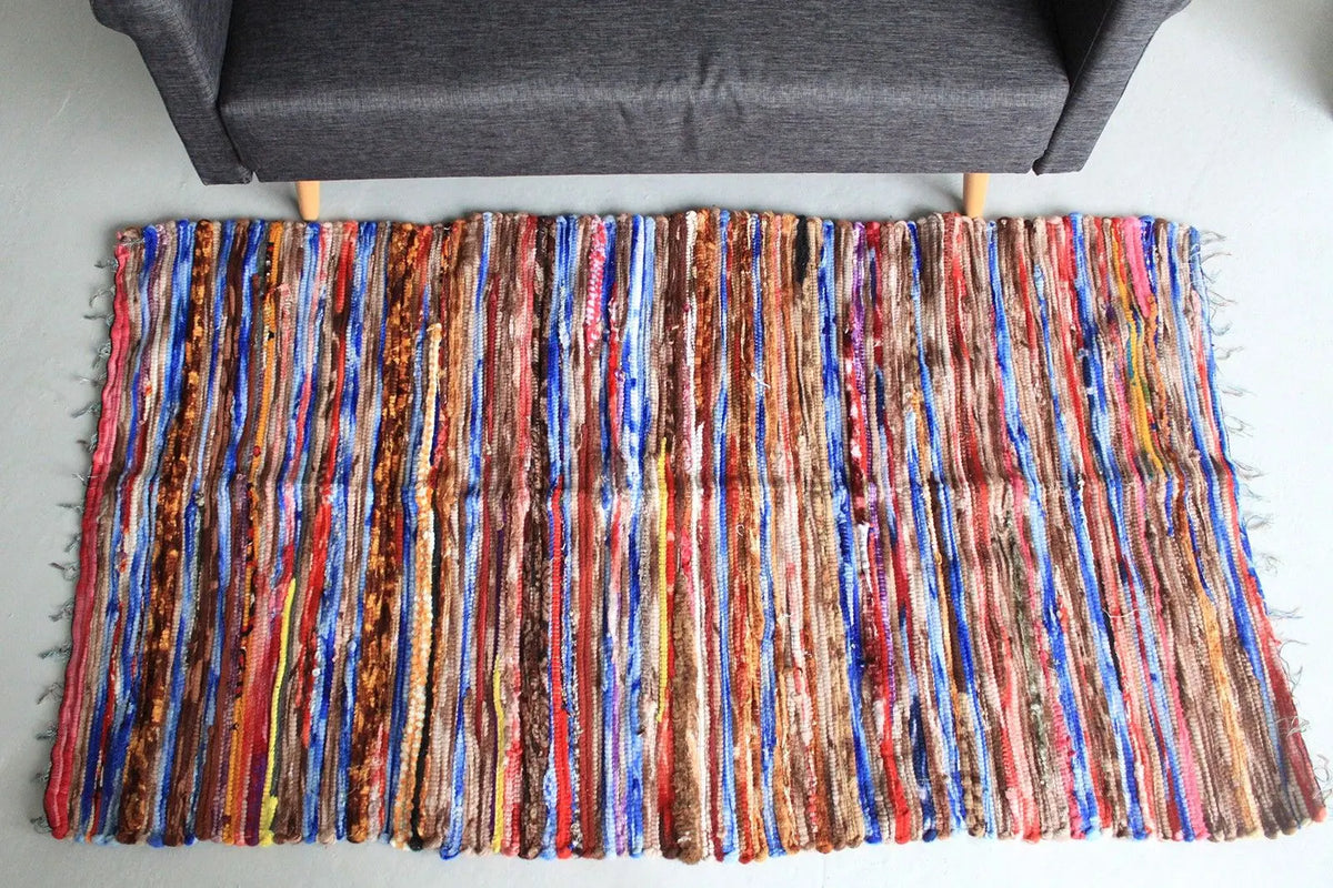 Alfombra de trapo india, 105x185cm, con tiras de tela multicolor en tonos cálidos y azules, estilo artesanal y casual