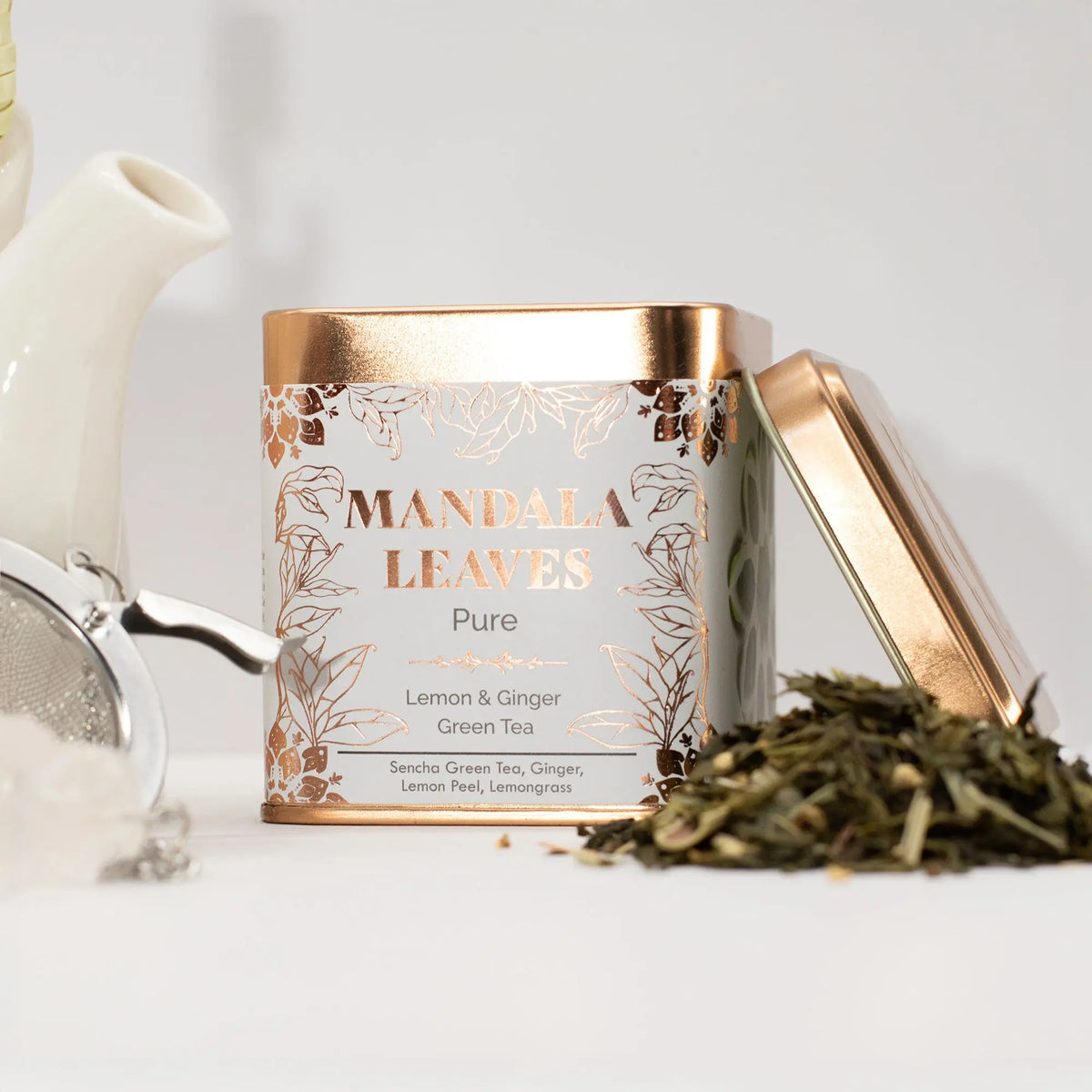 Lata cuadrada blanca y bronce con té verde de limón y jengibre, junto a un infusor y hojas de té sobre fondo claro.