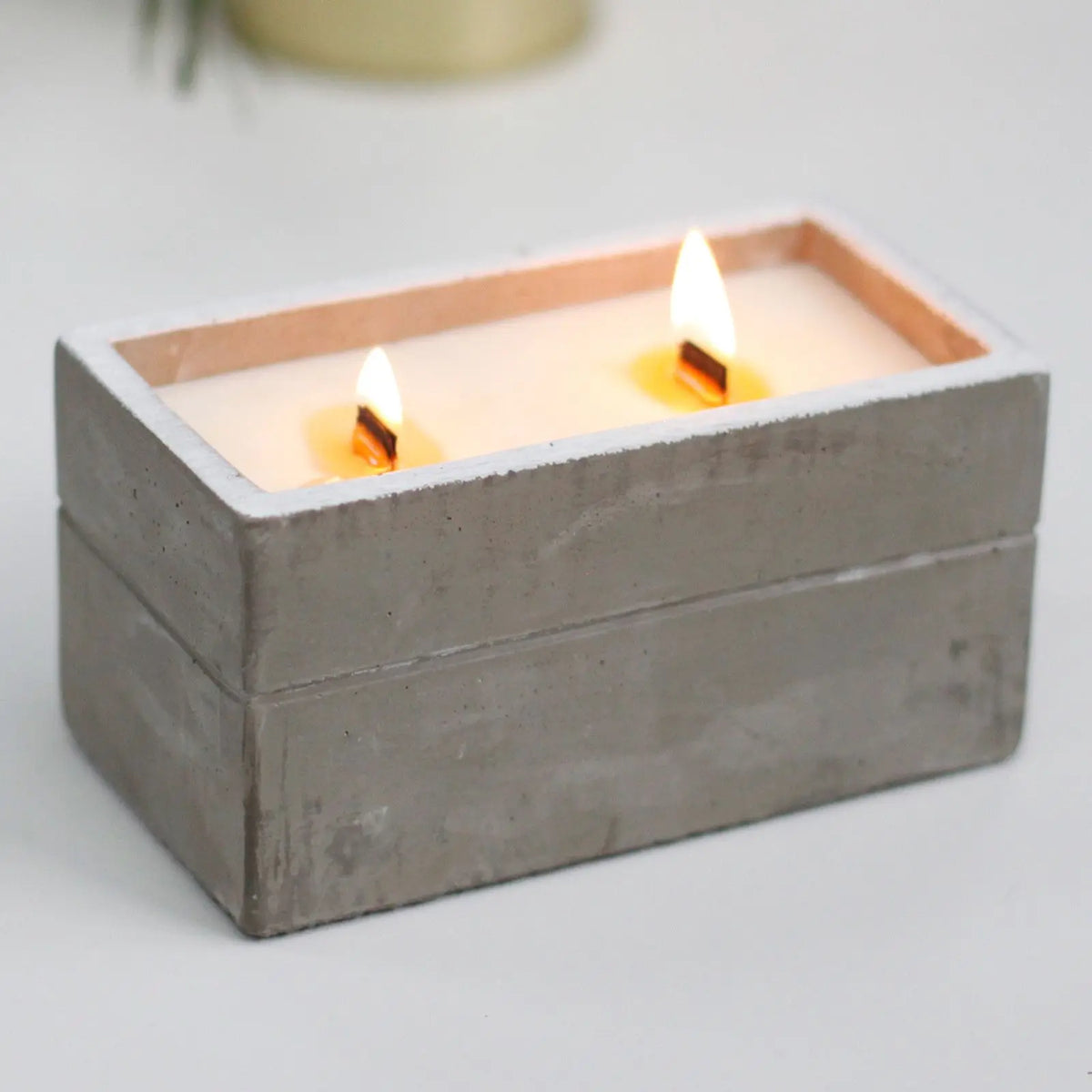 Vela rectangular de hormigón gris con dos mechas de madera encendidas y cera blanca en un recipiente de estilo minimalista