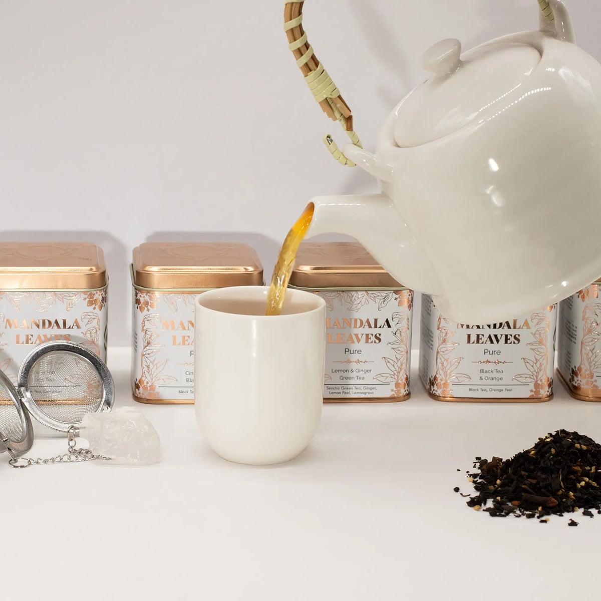Tetera blanca sirviendo té en una taza, rodeada de latas metálicas doradas, un infusor y té suelto sobre fondo blanco.