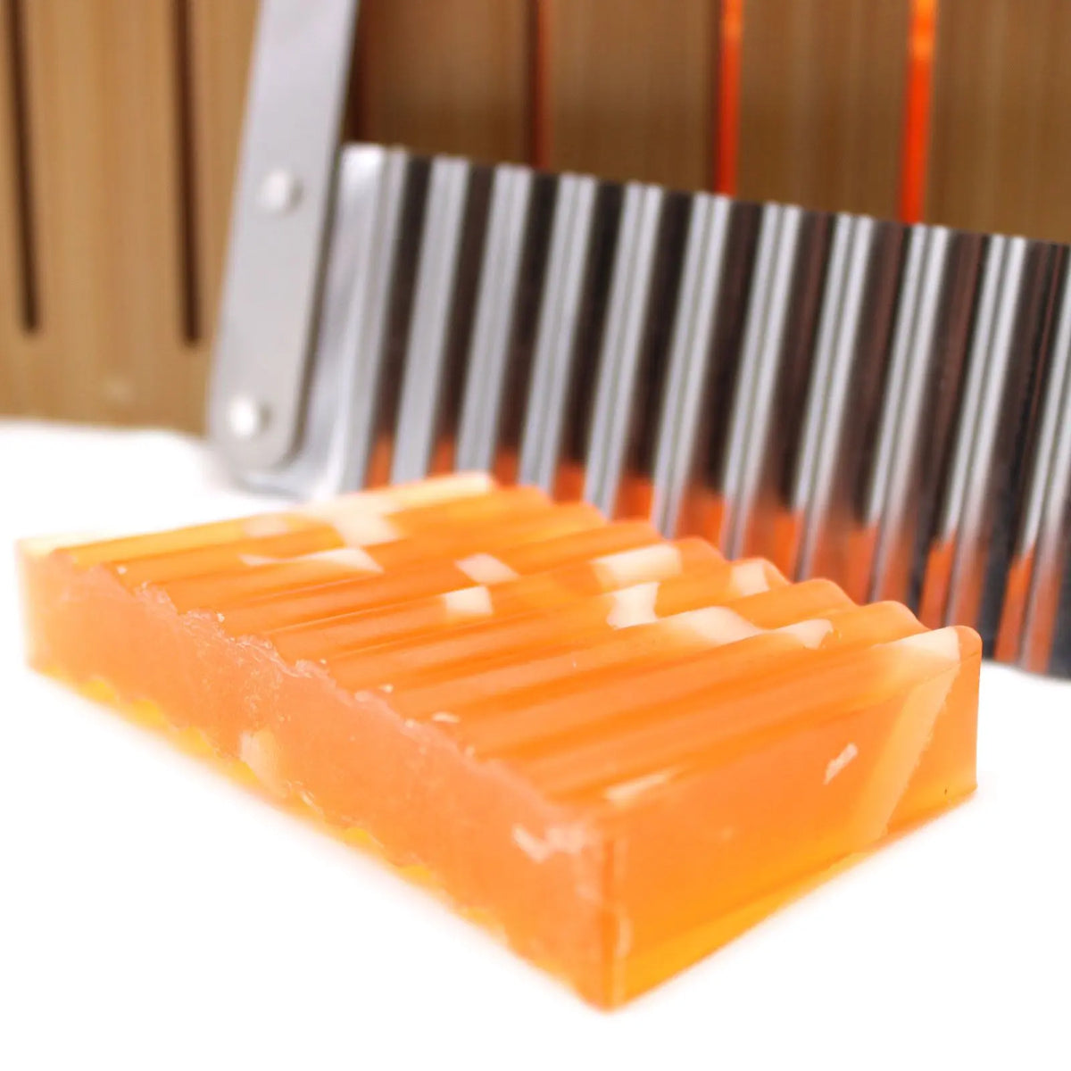 Cortador de jabón de acero ondulado y base de madera con una pastilla de jabón naranja texturizada al frente.