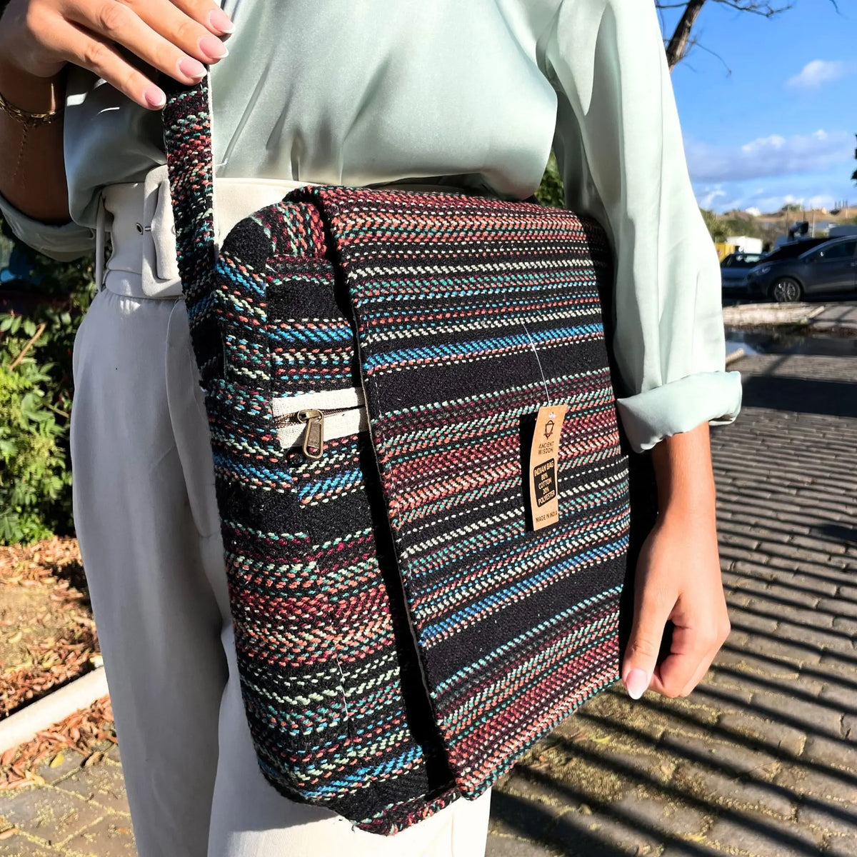Bolso bandolera de tela tejida con rayas multicolores sobre fondo negro, estilo rústico con cierre de cremallera.