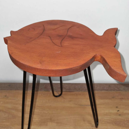 Soporte para plantas con forma de pez de madera terracota y patas metálicas negras sobre superficie de madera.