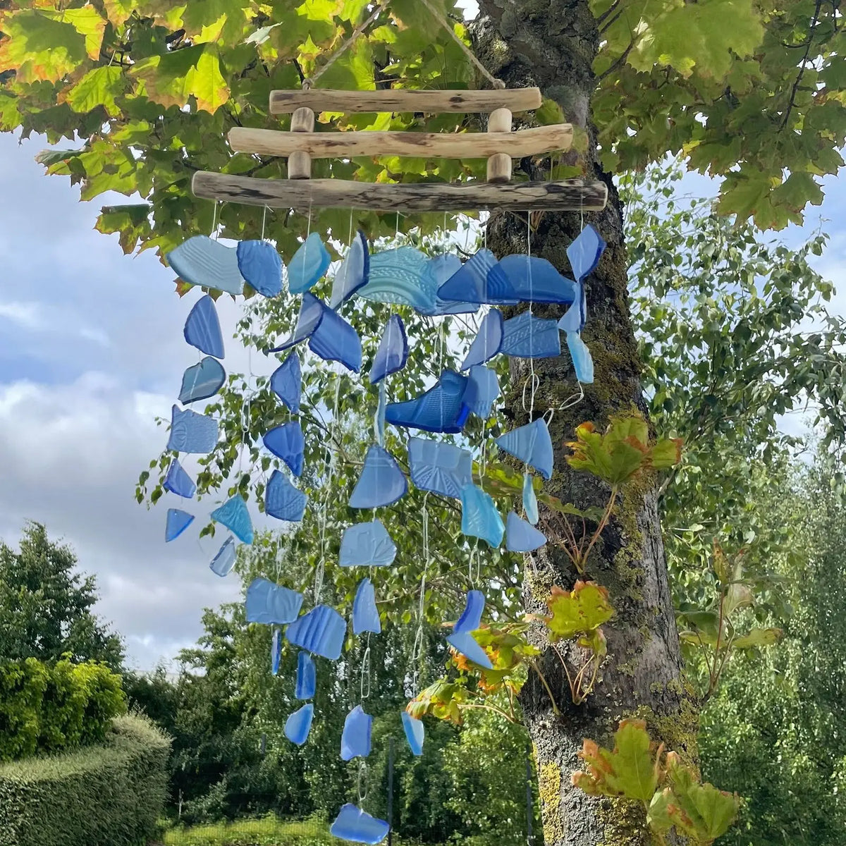 Carrillón artesanal con piezas de vidrio reciclado azul, colgado de una estructura de madera natural en un árbol.