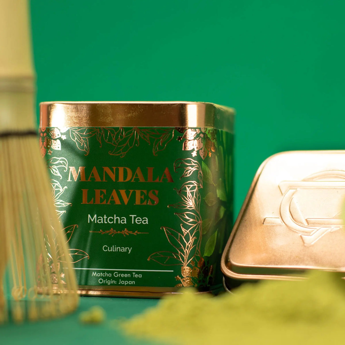 Lata cuadrada verde de té matcha con detalles dorados, un batidor de bambú y polvo de matcha sobre fondo verde.